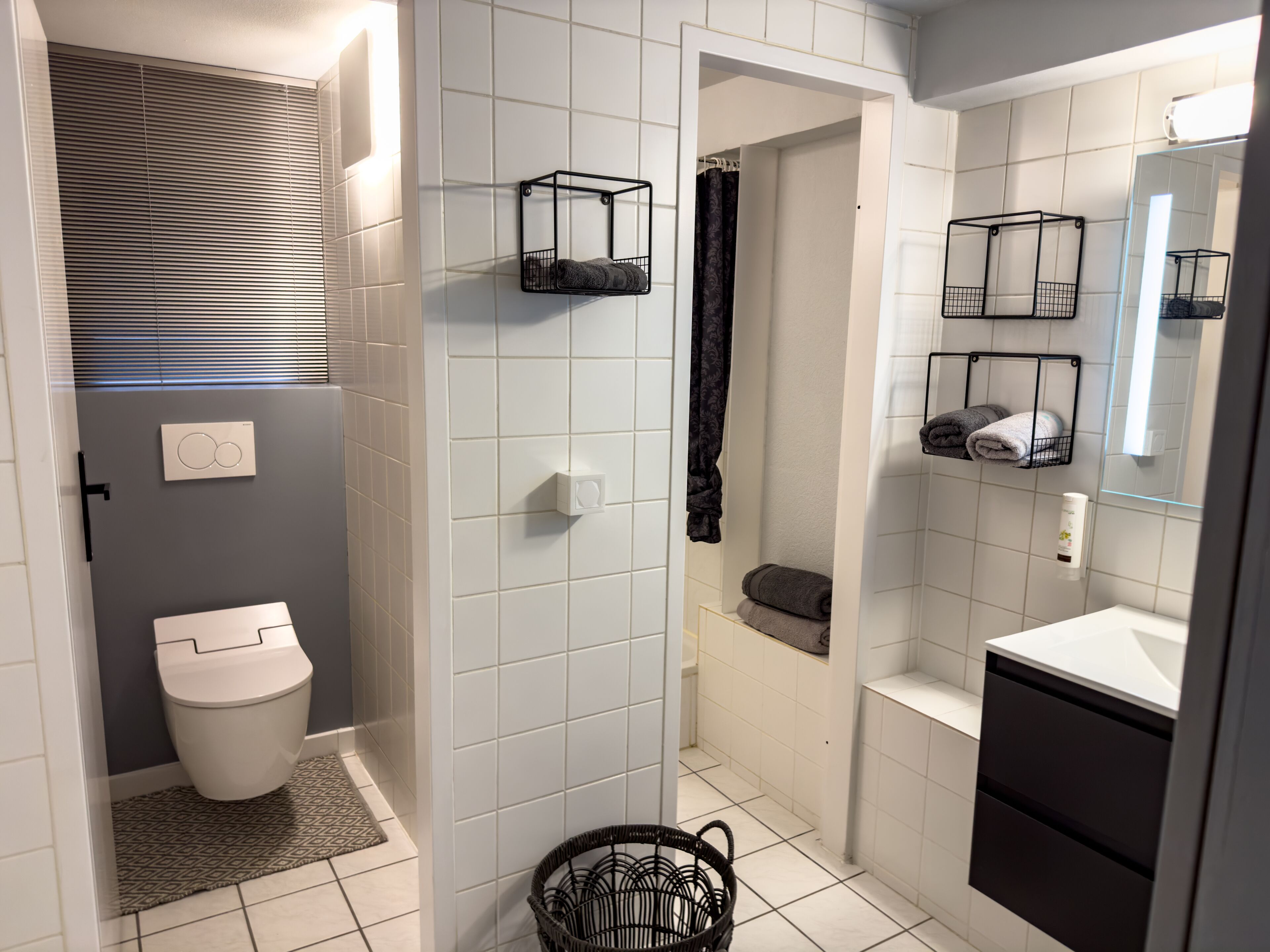 Ein modernes Badezimmer mit weißen Fliesen, einer Toilette, einer Dusche und einem Waschbecken mit Spiegel.
