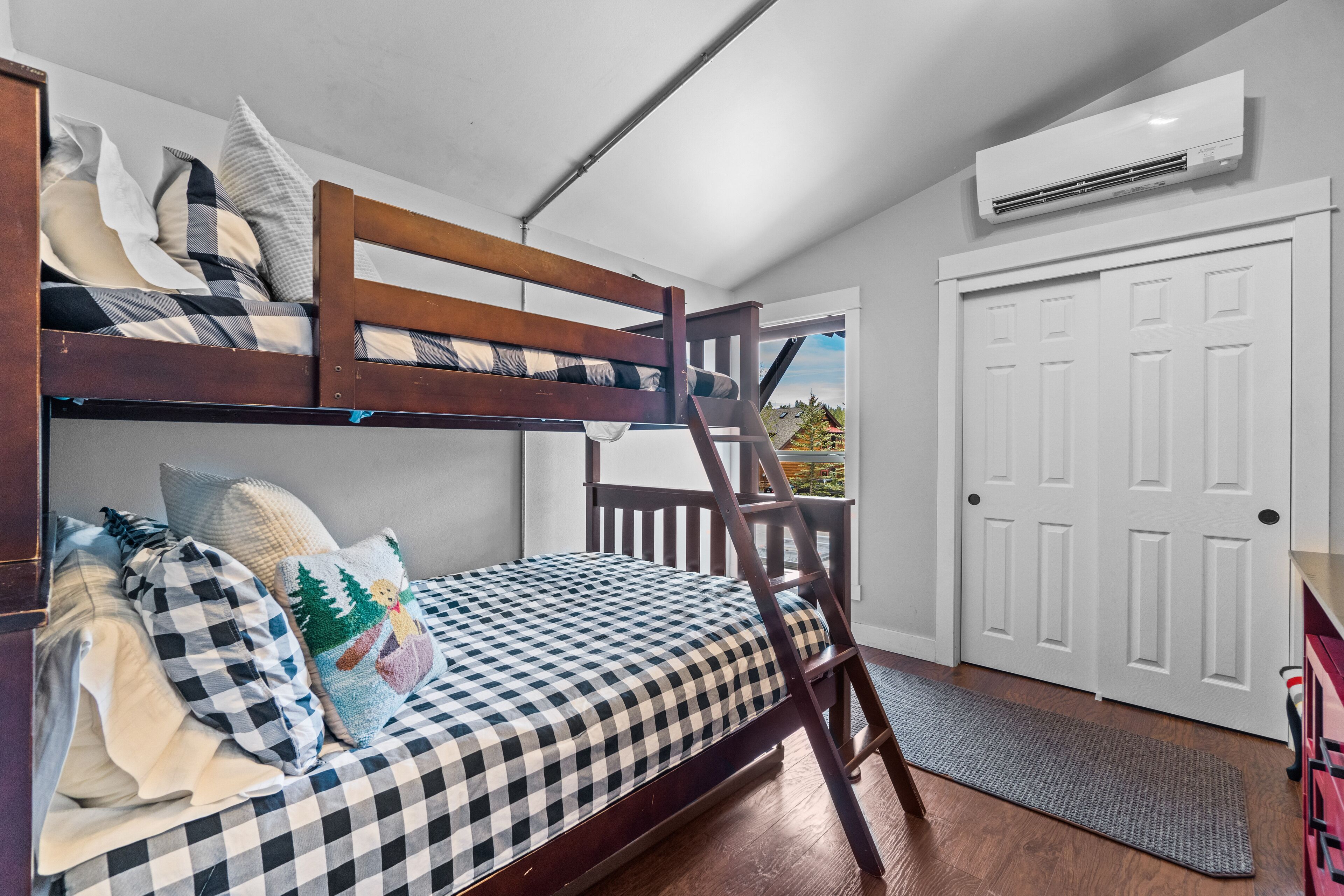 A bunk bed with a checkered mattress, pillows, and a ladder. There is a white door and an air conditioning unit on the wall.