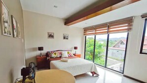 A bedroom with a bed, bedside tables, a fan, a round table, and a large window with a view of greenery and buildings.