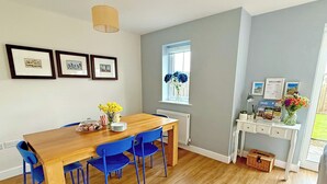 A dining area with a wooden table, blue chairs, framed pictures on the wall, a window with flowers, and a reception desk with a welcome sign. - 3 bedroom accommodation in Buxton (Buxton)