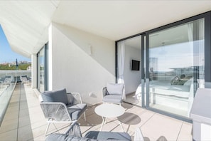Ein moderner Balkon mit weißer Möbel, eine Glastür, die zu einer Terrasse führt, und ein freier Blick auf die Stadtlandschaft.