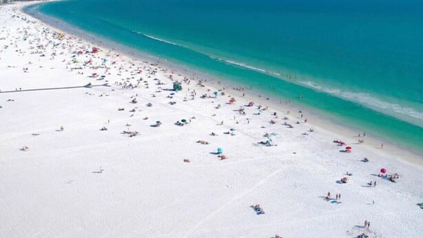 Een strand met vele parasols en mensen, een heldere blauwe zee en een breed zandstrand.