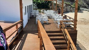 A wooden deck with tables and chairs, leading to a beach area with a wooden pergola overhead. - Paradise By The Sea - TheWhaleBeachHouse (Atouguia da Baleia)