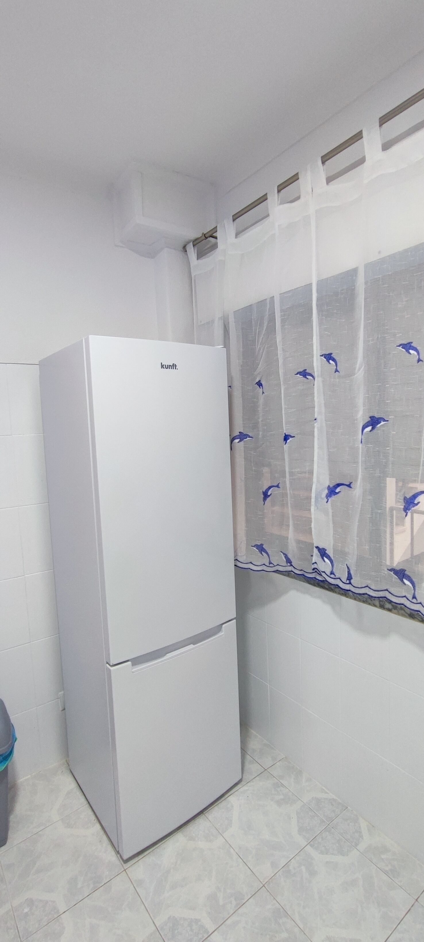 A white refrigerator in a corner of a room with tiled flooring and a window with a dolphin pattern curtain.