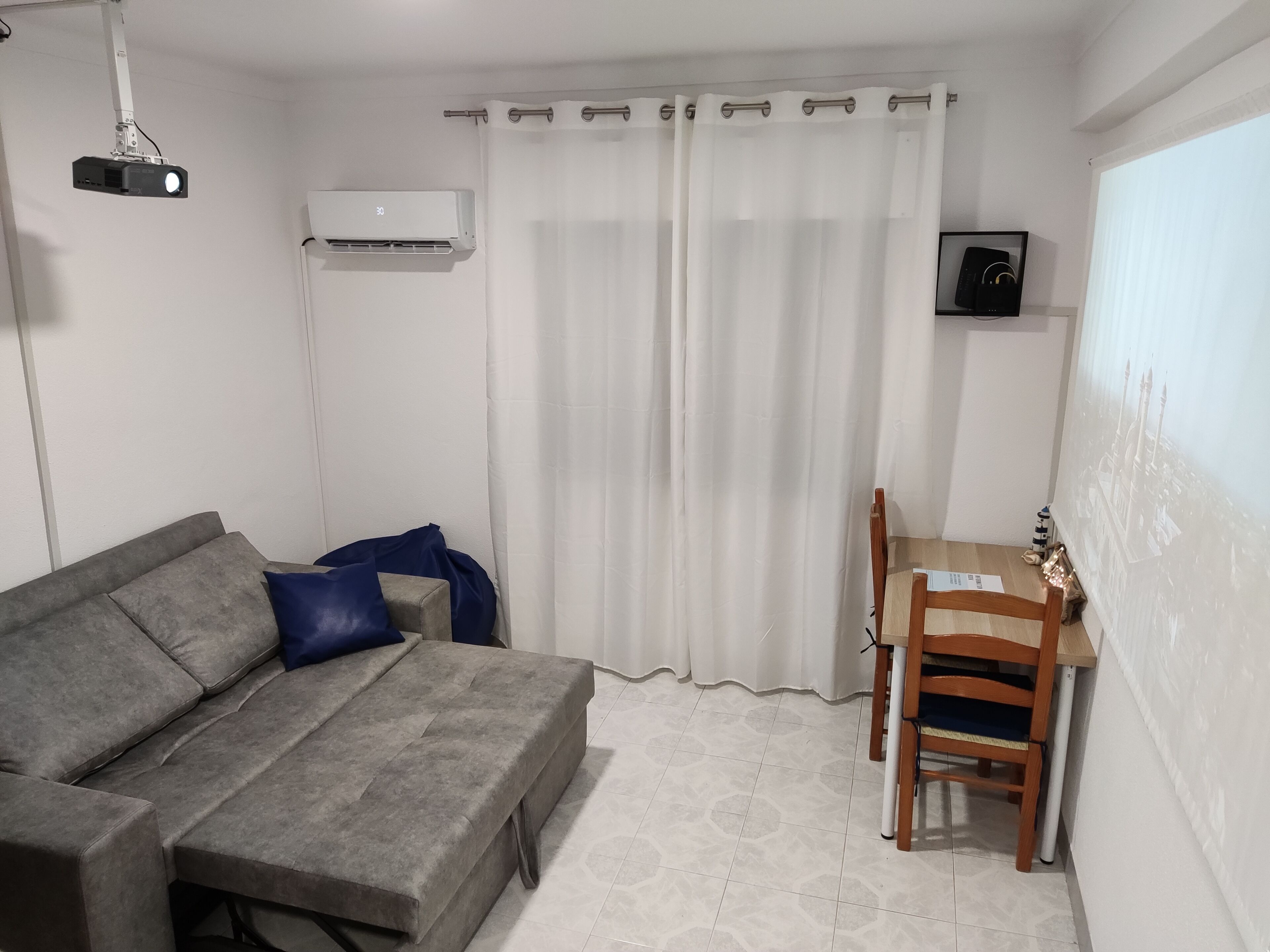 A small, modern room with a grey sofa, a wooden chair, a white curtain, and a projector mounted on the ceiling.