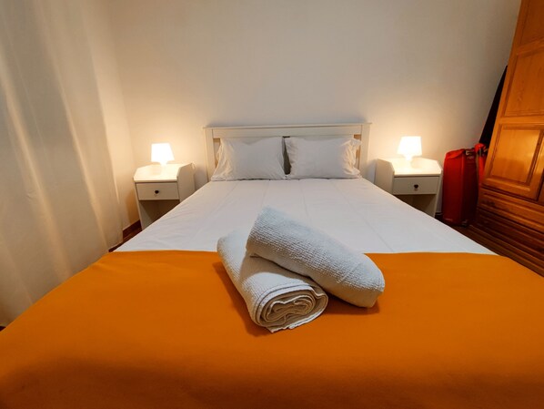 A neatly made bed with white linens and a rolled towel, two bedside lamps, and a wooden wardrobe.
