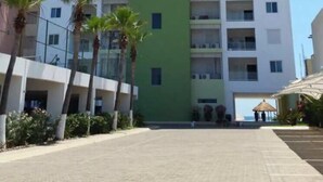 A multi-story hotel with a central green section, white and pink buildings on either side, and a paved walkway with palm trees. - Condos Ventana R (Mazatlán)