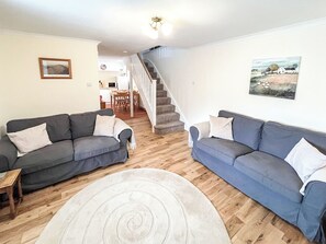 Una sala de estar con dos sofás, una alfombra, una escalera y un comedor al fondo.