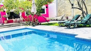 Una piscina rectangular con agua azul cristalina, rodeada por un solárium con baldosas. Hay sillas de descanso y una pequeña mesa con sillas cerca.