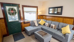 A living room with a sectional sofa, two ottomans, a wooden tray, a green door, and a window with a view of parked cars. - Stay near Mirror Lake, enjoy our cozy 2-bedroom cottage (Lake Placid)