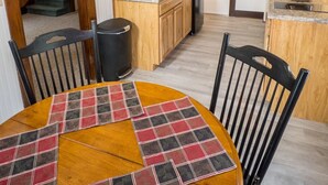 A dining area with a round table covered by a plaid tablecloth, wooden chairs, and a kitchen in the background. - Stay near Mirror Lake, enjoy our cozy 2-bedroom cottage (Lake Placid)
