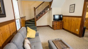A living room with a grey sectional sofa, a wooden coffee table, a staircase, a television on a wooden stand, and a framed picture on the wall. - Stay near Mirror Lake, enjoy our cozy 2-bedroom cottage (Lake Placid)