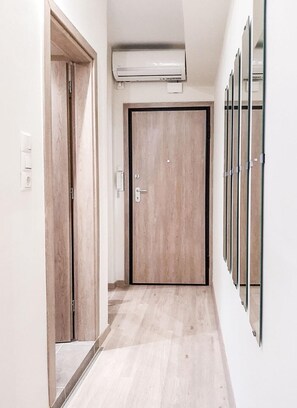 Un couloir avec une porte en bois, un appareil de climatisation placé au-dessus, et un miroir fixé au mur sur le côté droit.