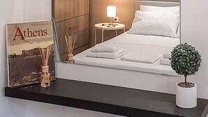A neatly made bed with white linens, a small bedside table with a lamp, a framed book titled 'Athens', a small potted plant, and a wooden wardrobe. - Byronico Acropolis Apartment by MPS (Αθήνα)