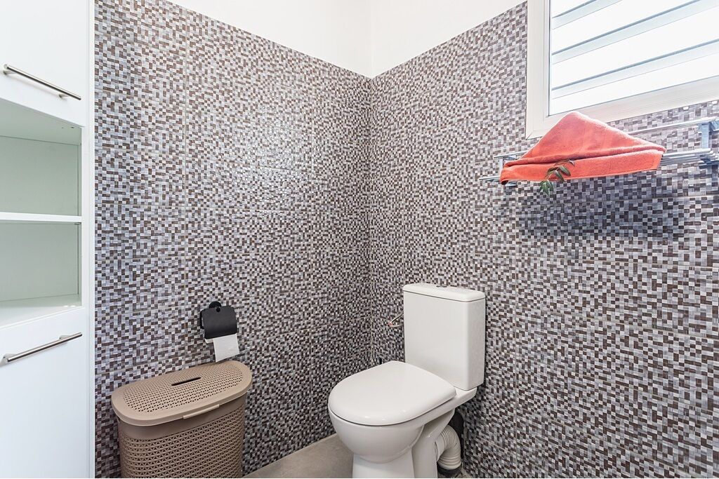 A small bathroom with a tiled wall, a toilet, a trash bin, and a towel rack with a towel.