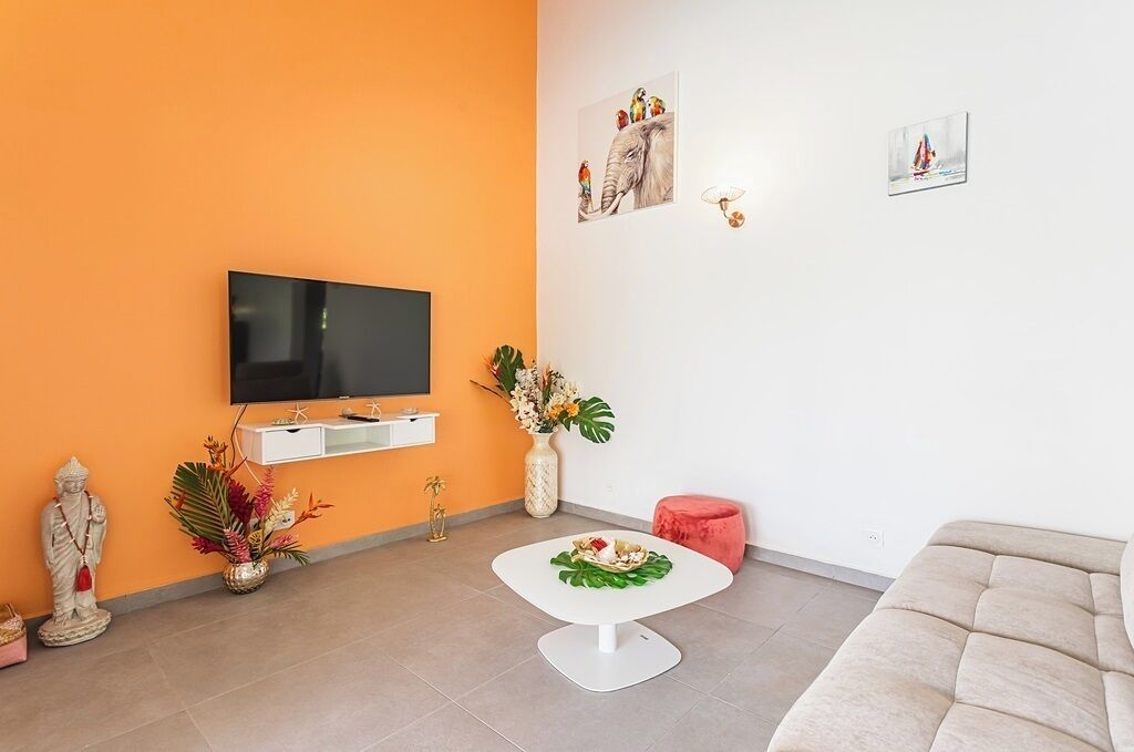 Una sala de estar con un televisor de pantalla plana, un sofá blanco, una mesita redonda con un plato, una estatua y un jarrón con flores.