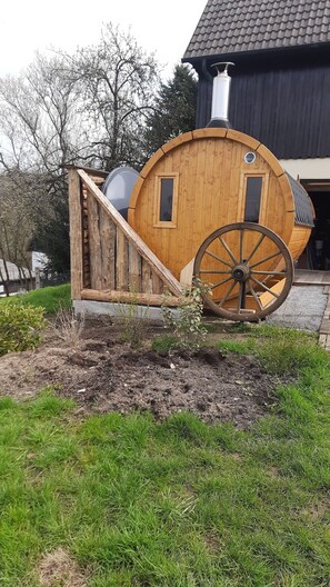 Une structure en bois ressemblant à un tonneau, dotée d’une roue, d’une fenêtre et d’une porte, située dans un jardin aménagé avec de la pelouse et des arbres.