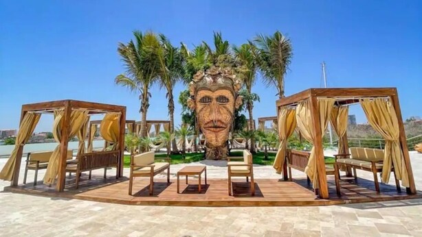 Un espace de détente aménagé sur la plage, avec des cabanes en bois et des sièges, des palmiers, et une grande sculpture en pierre en arrière-plan.