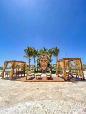 A beachside lounge area with wooden cabanas and seating, palm trees, and a large stone sculpture in the background. - Condos Marina PP (Mazatlán)
