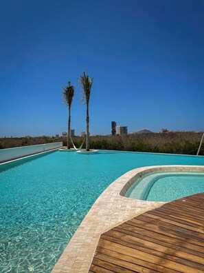 Una piscina azul cristalina con un deck de madera, rodeada de palmeras y un paisaje urbano al fondo.