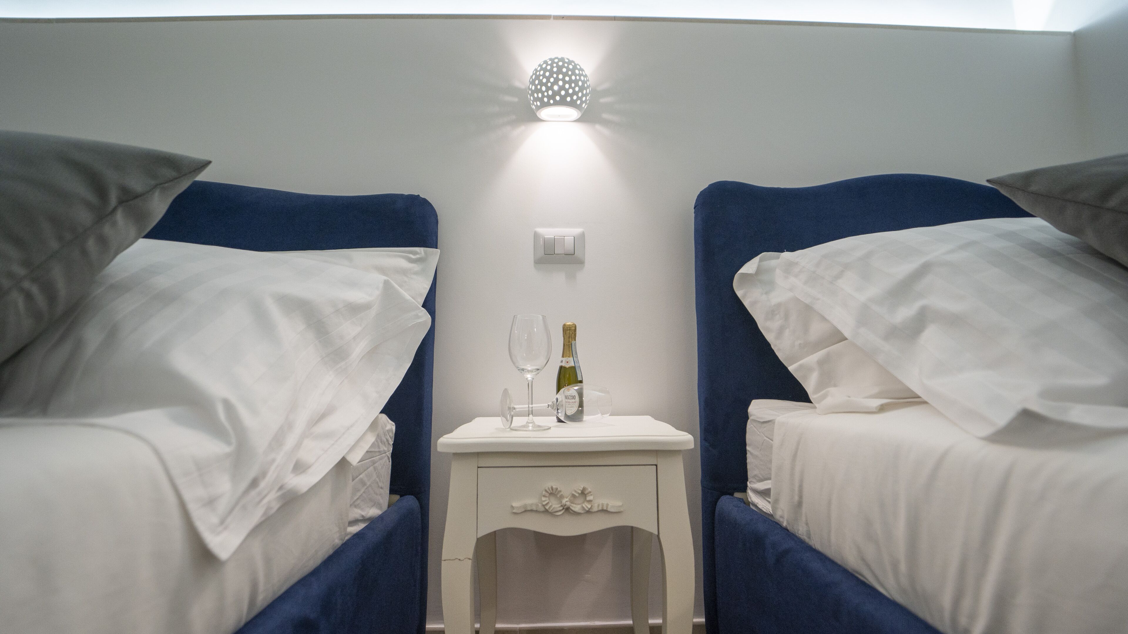 A hotel room with two beds, each with a blue headboard and white bedding. There is a small bedside table between the beds with a bottle of champagne and a glass.