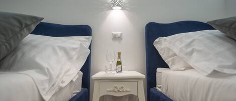 A hotel room with two beds, each with a blue headboard and white bedding. There is a small bedside table between the beds with a bottle of champagne and a glass.