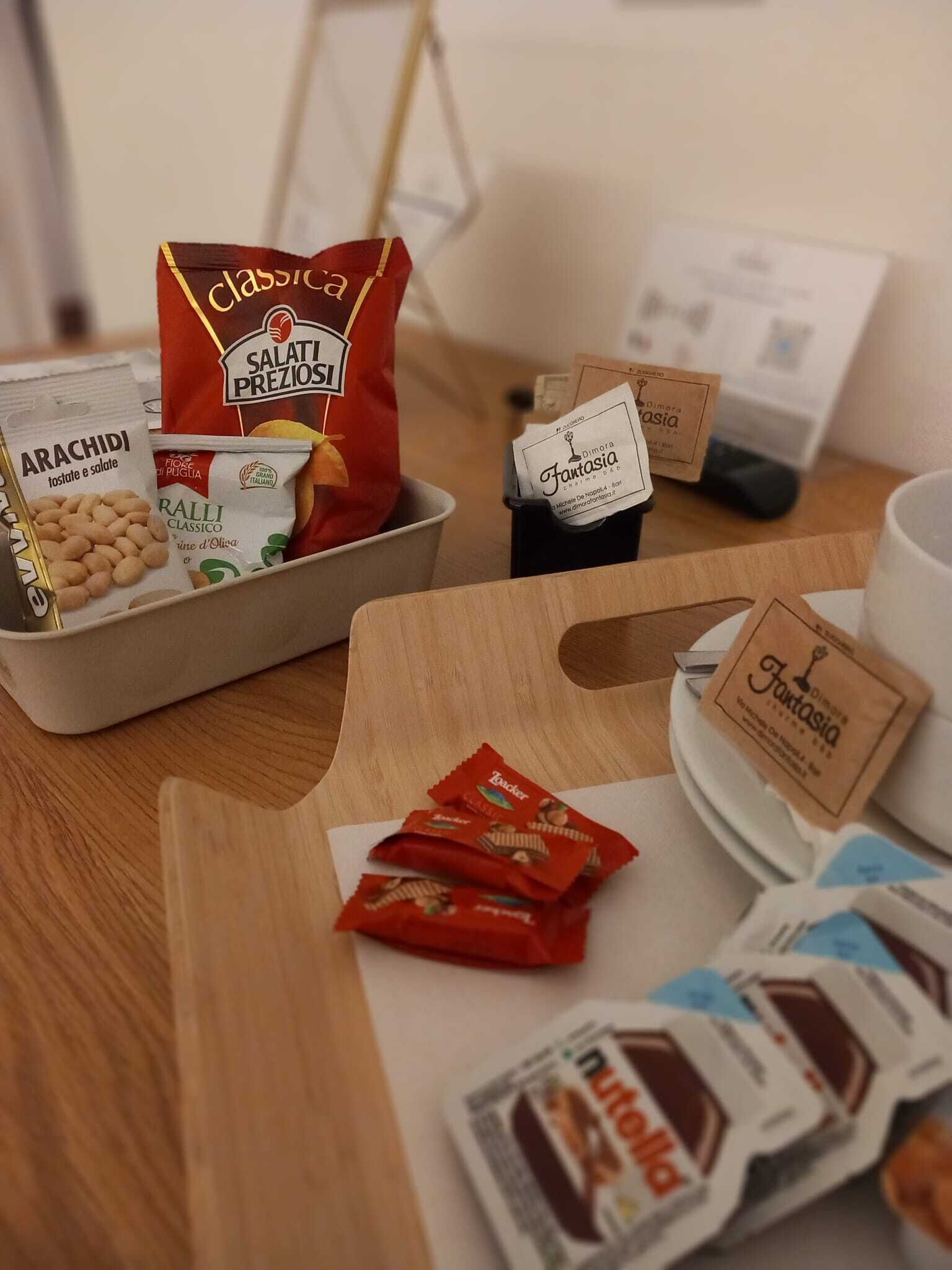 A wooden table with food items including a bag of salami, a packet of arachidi, and a packet of ralli.