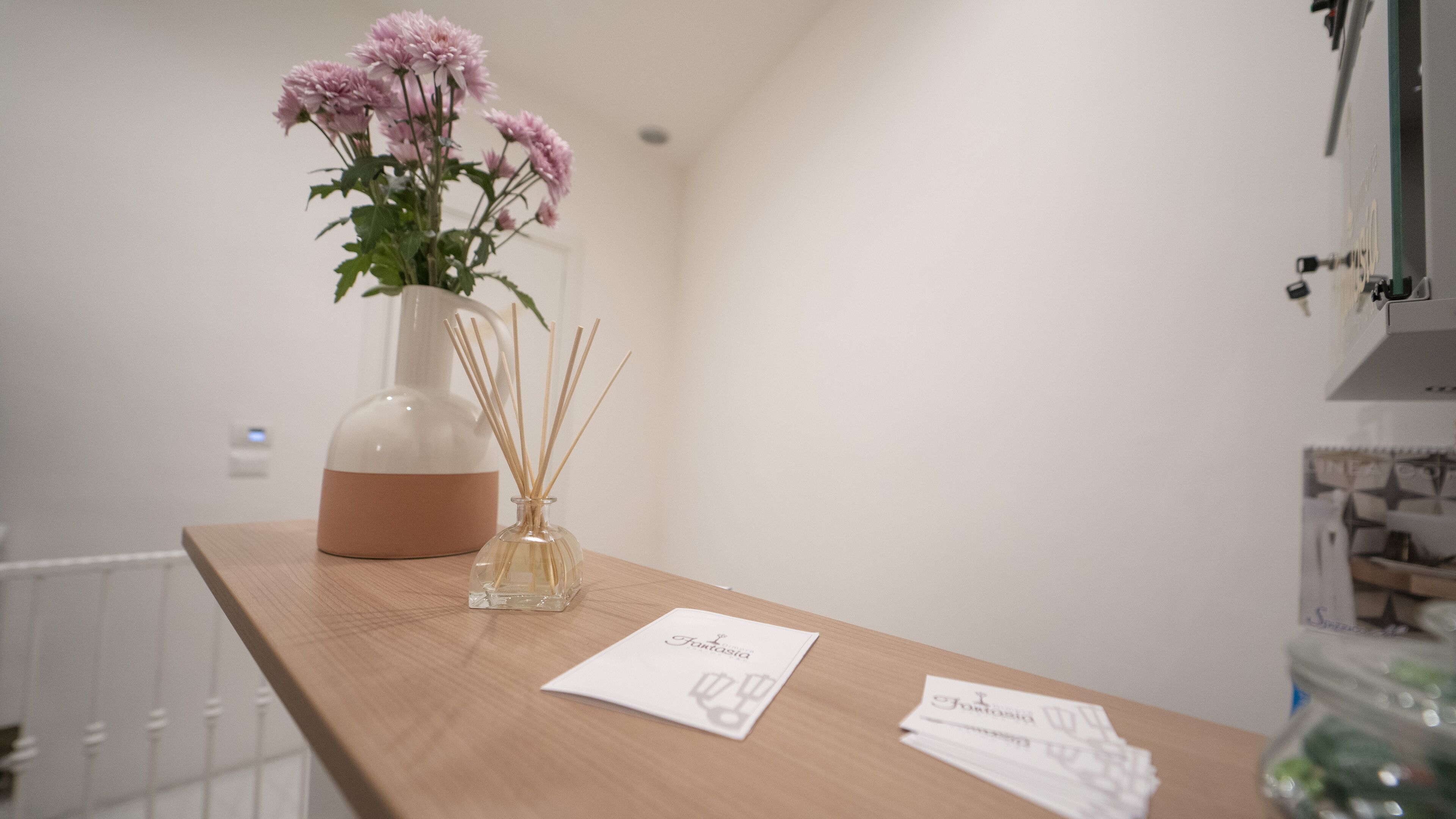 Um balcão de madeira com um vaso de flores cor-de-rosa, um difusor e um cartão de visita.