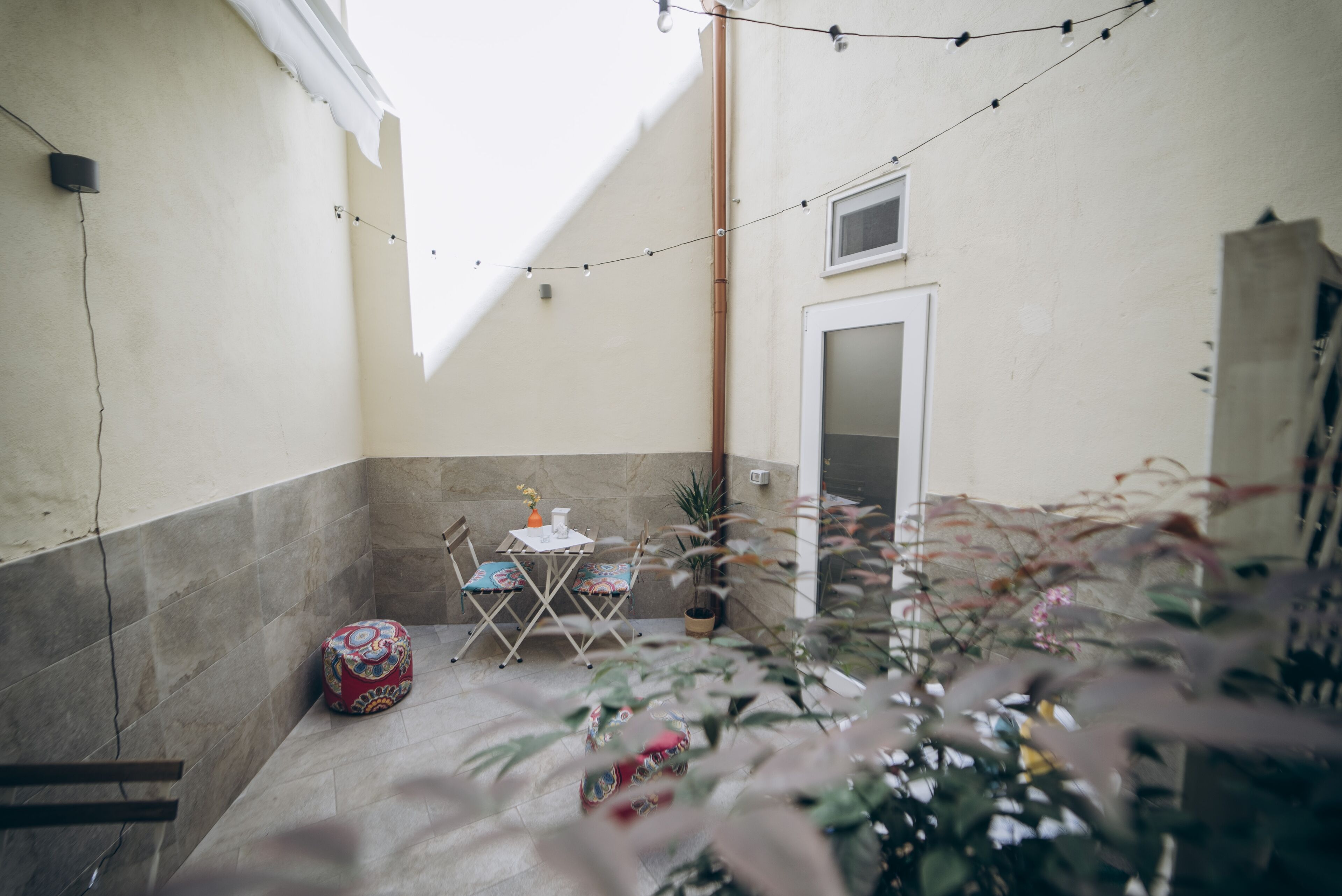 Un pequeño patio amurallado con una mesa, sillas, una planta en maceta y luces colgantes.