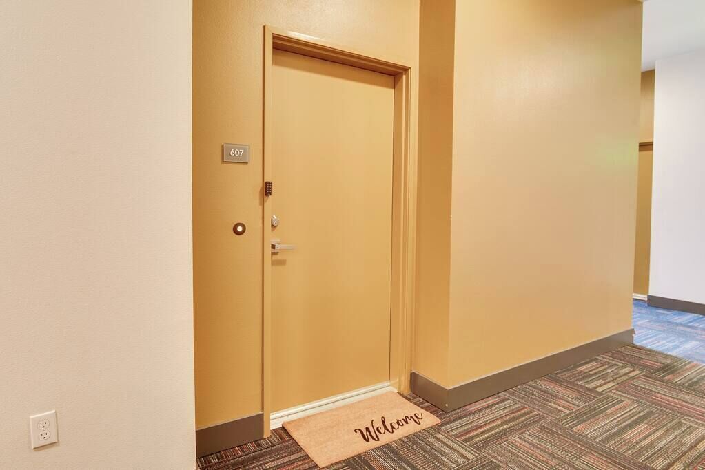 L’entrée d’une chambre d’hôtel, avec une porte fermée, un paillasson et un sol recouvert de moquette.