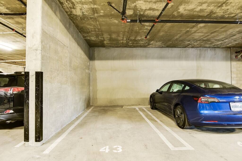 Un parking avec des murs en béton, une voiture bleue garée dans une place marquée du numéro 43, et des tuyaux apparents au plafond.