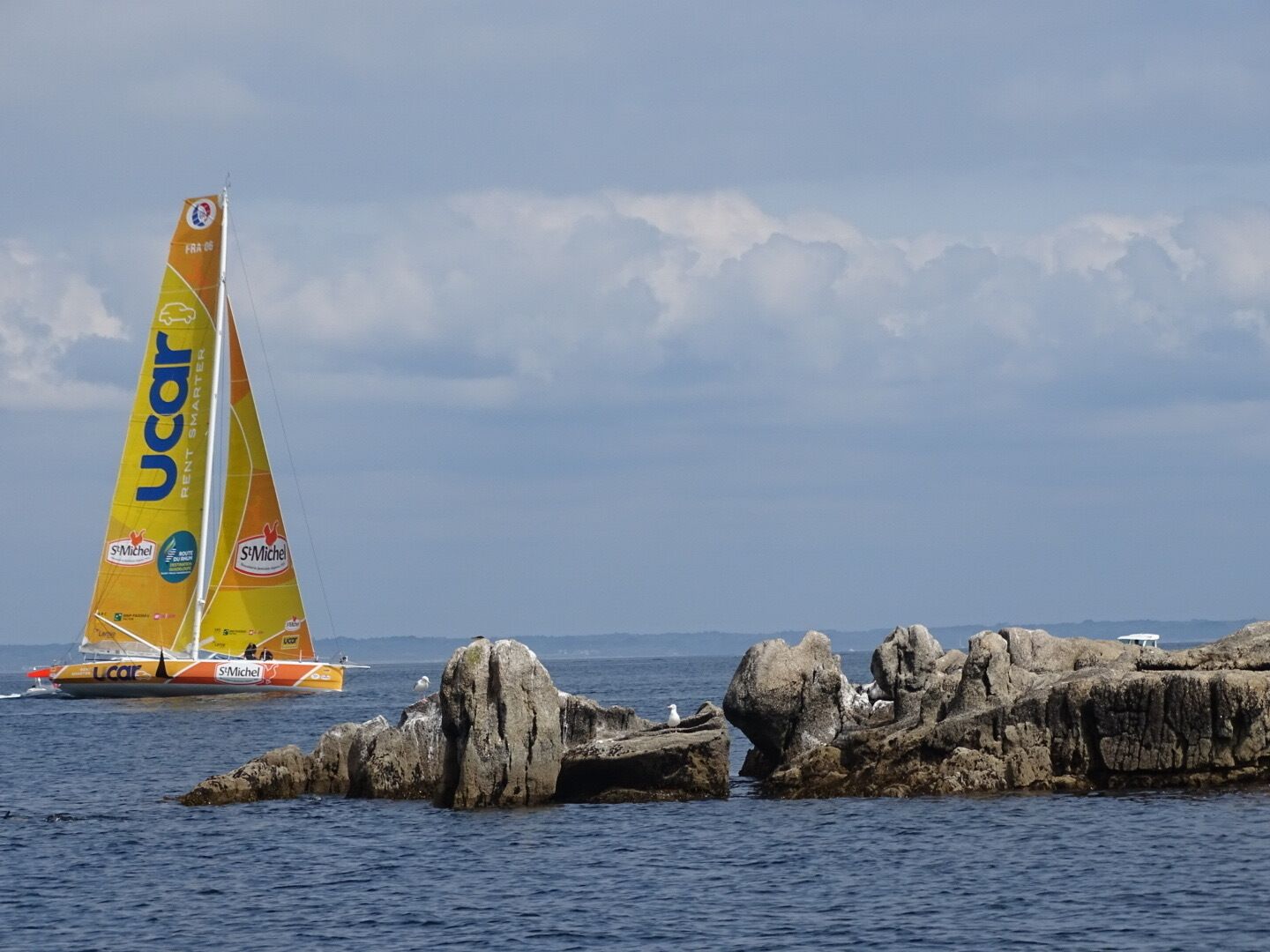 A sailboat with a yellow and orange sail, sailing near rocky formations in the water.