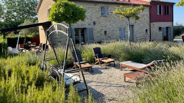 Een stenen gebouw met een rood dak, een terras met stoelen en een tafel, en een schommelset.
