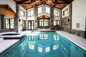 Una amplia piscina interior con paredes de piedra y vigas de madera, rodeada de grandes ventanales.