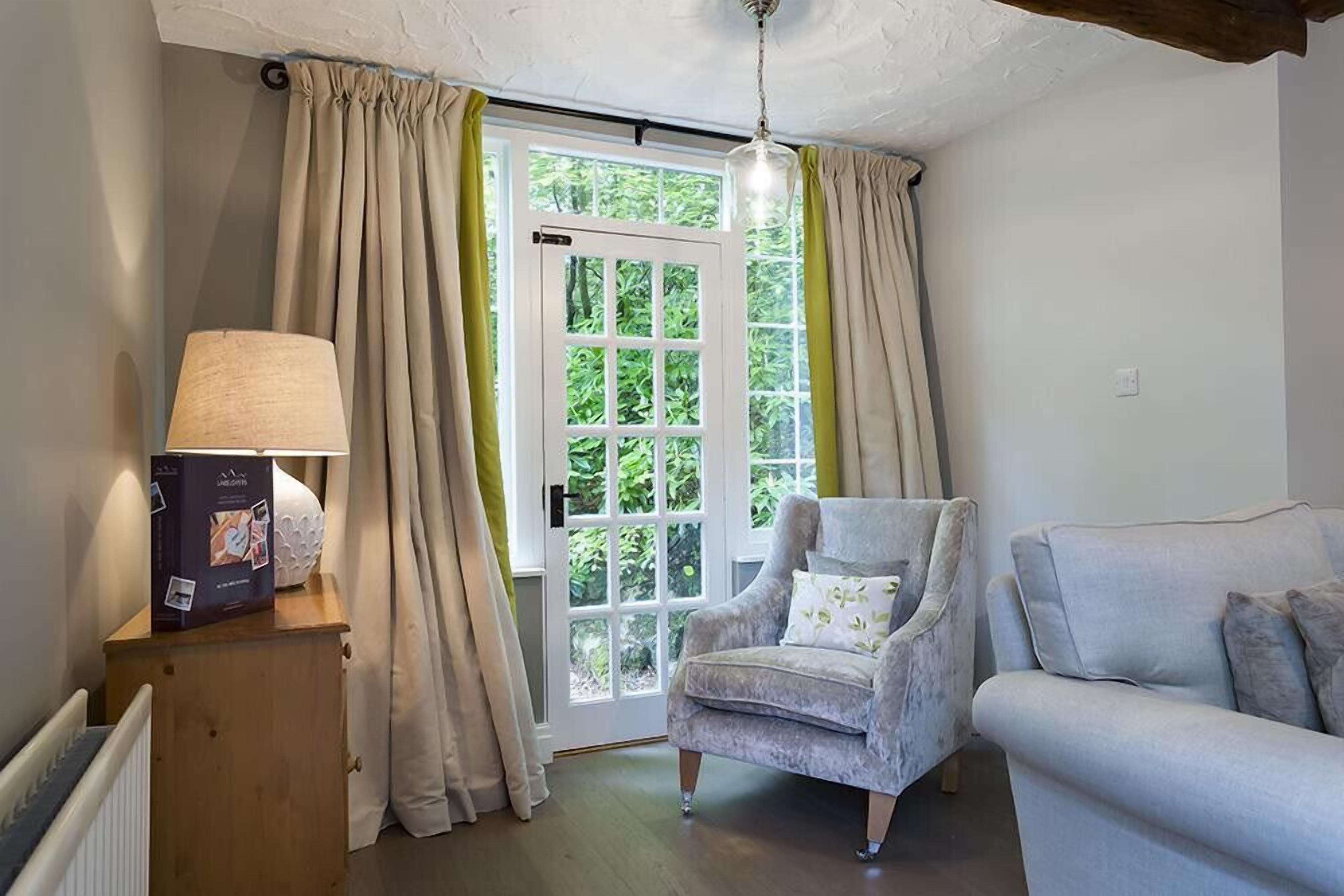 Una habitació acollidora amb un armari de fusta, una làmpada, un armchair gris amb un coixí i un sofà. Un gran finestral amb cortines deixa entrar llum natural.