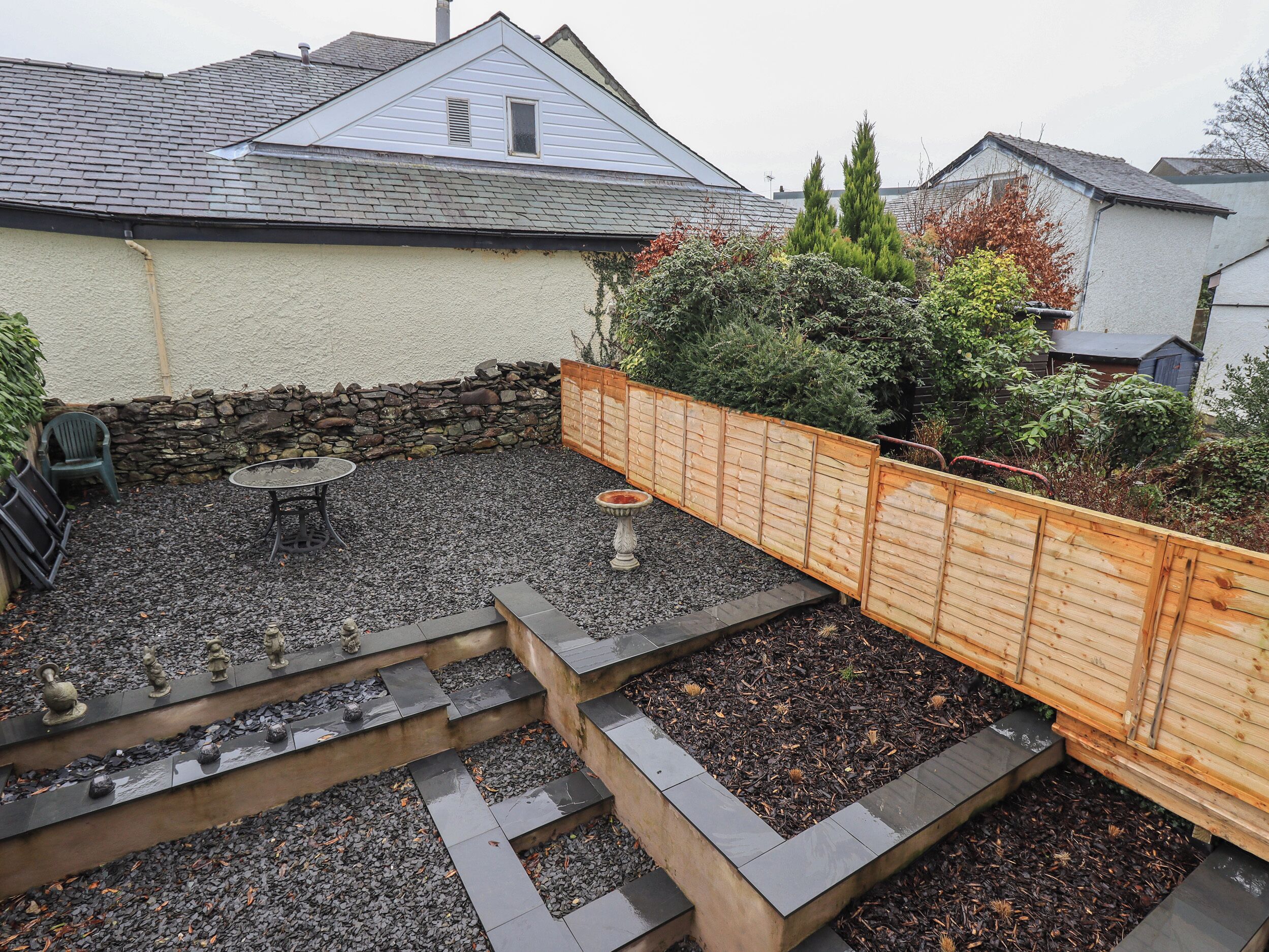 A backyard with a stone wall, a wooden fence, and a garden area with steps leading to a table and chairs.