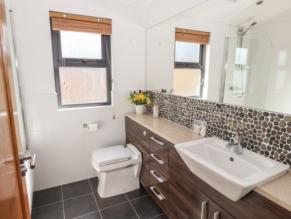 Baño con inodoro, lavamanos y ducha. Cuenta con dos ventanas con persianas, un espejo y un florero decorativo con flores.