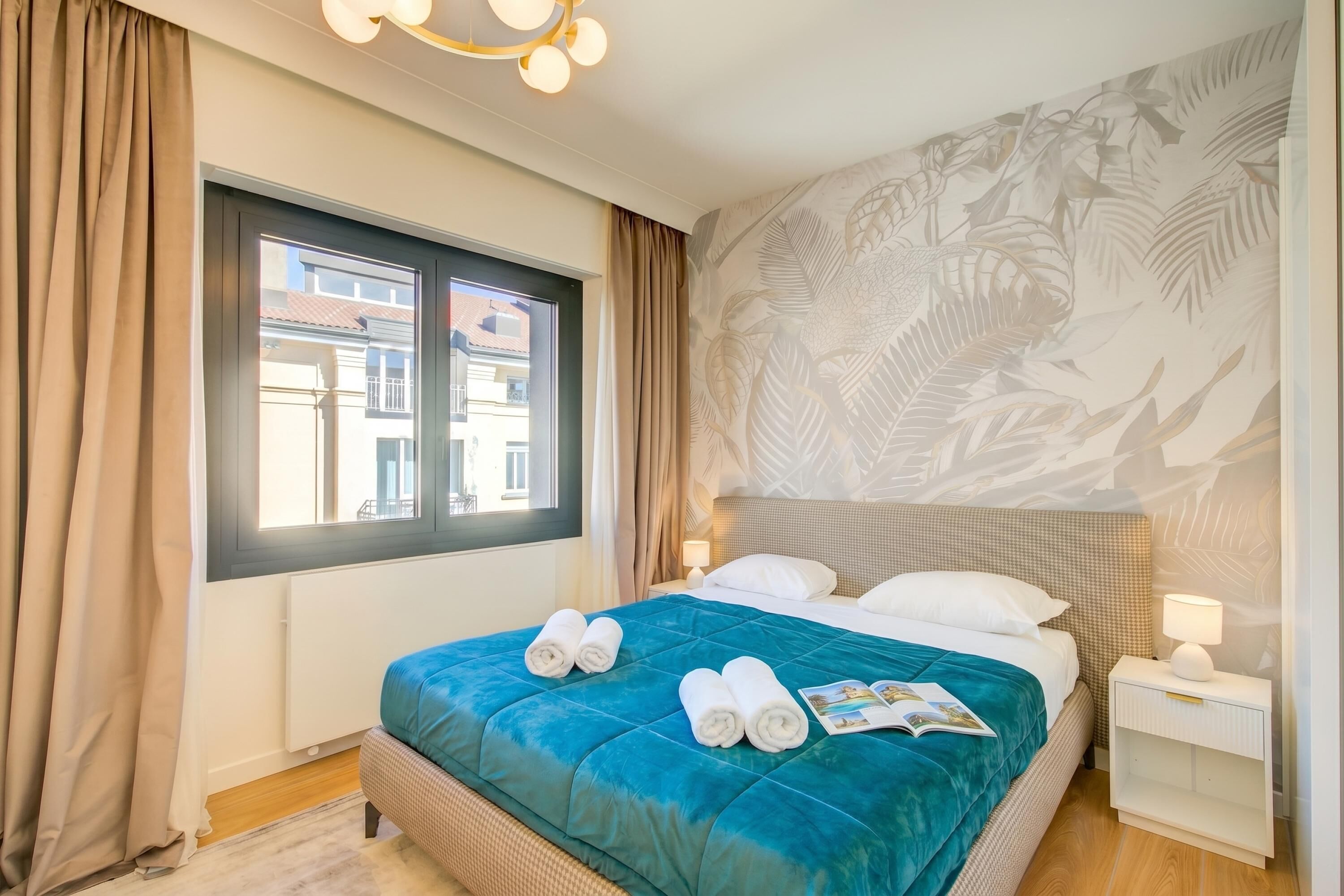 Quarto de hotel com cama, janela, mesa de cabeceira e parede com padrão de folhas.
