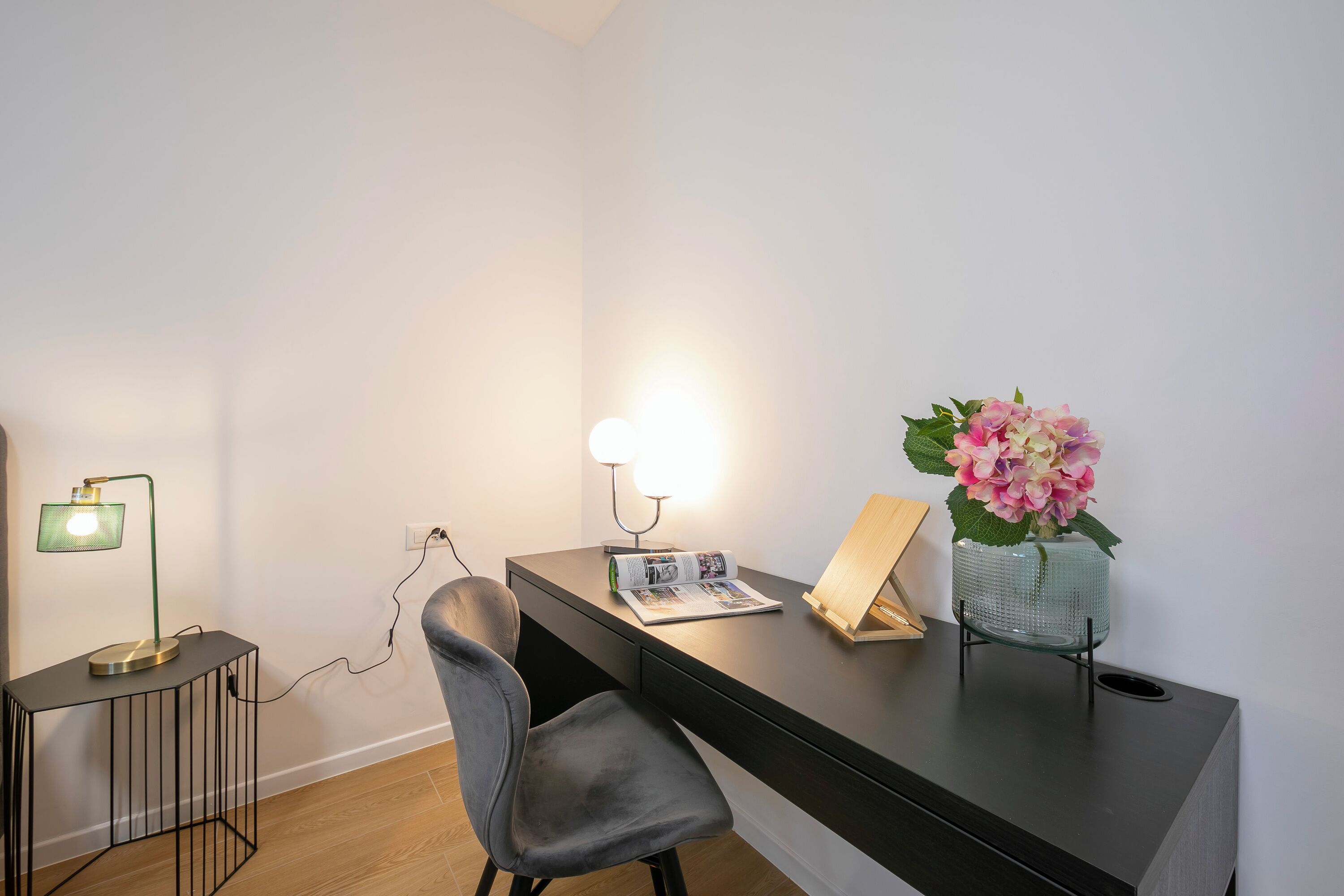 Een moderne werkruimte met een bureau, stoel, lamp, tijdschrift en een vaas met bloemen.