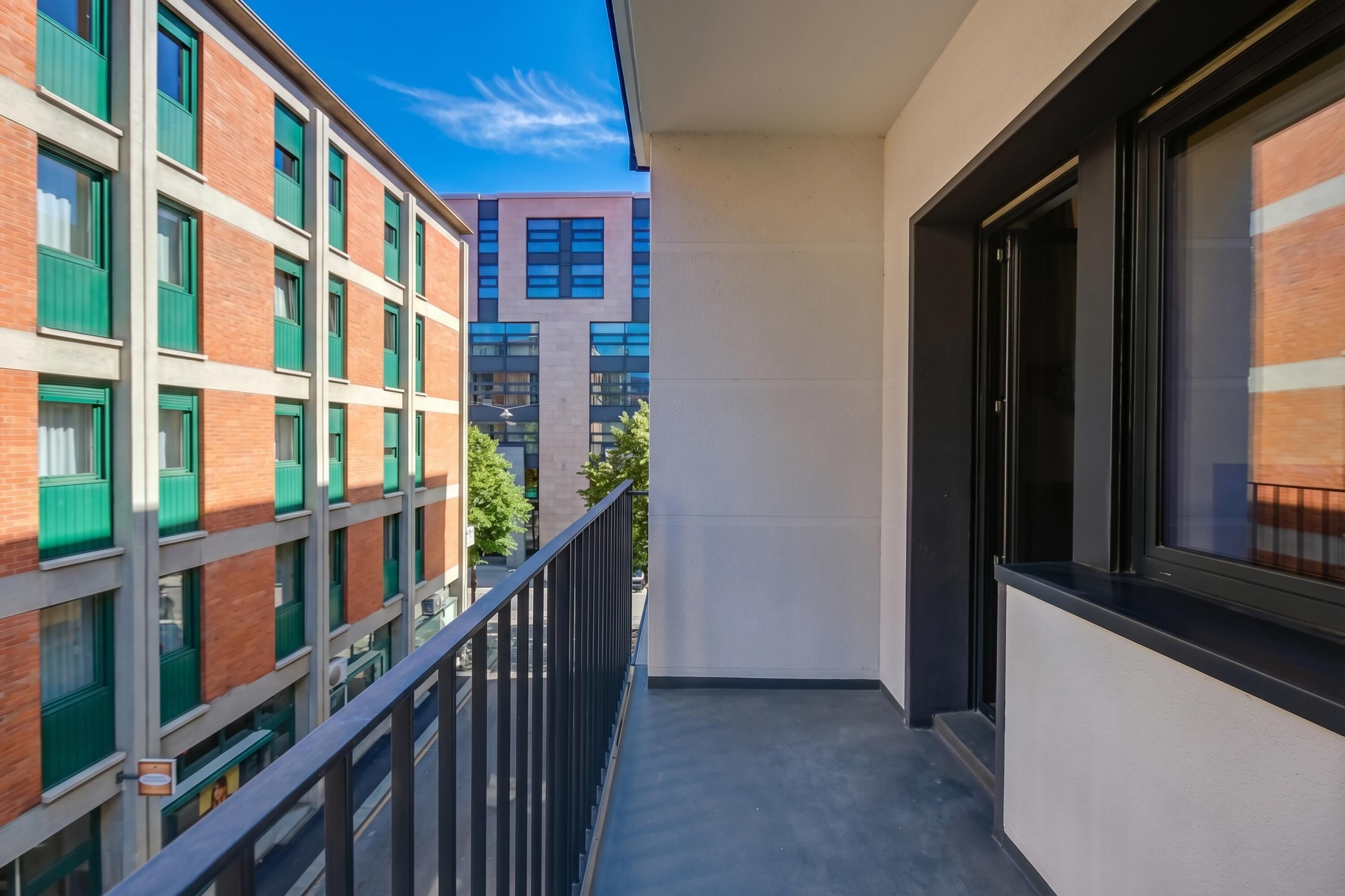 Un balcon étroit avec une rambarde noire, une vue sur les immeubles voisins et un ciel dégagé.