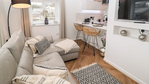 Ein modernes Wohnzimmer mit einer Couch, einer Essecke mit zwei Barhockern, einer Küche mit Fernseher und einem großen Fenster mit Vorhängen.