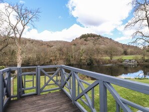 A wooden deck with a railing overlooking a calm lake, surrounded by trees and a hill in the background. - Riverside Retreat (Ulverston)