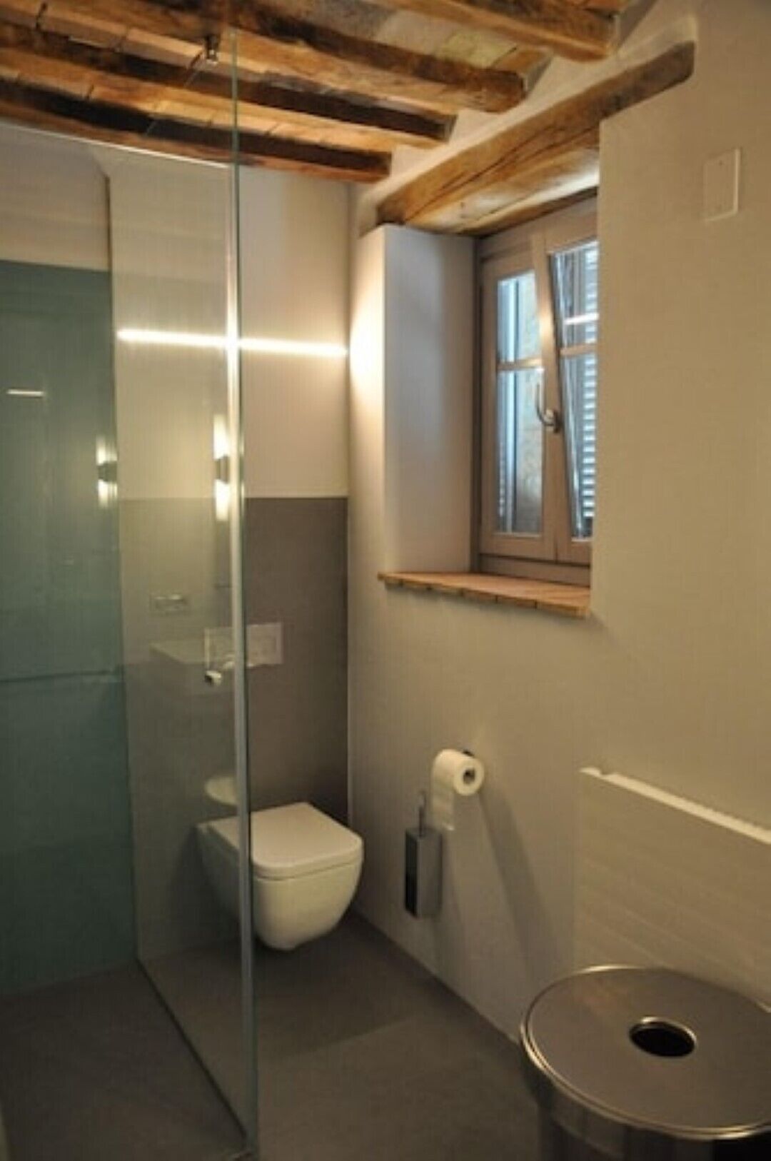 Une salle de bain avec une douche à parois de verre, des toilettes et une fenêtre dotée d’une étagère en bois.