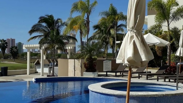 A swimming pool with lounge chairs and umbrellas, surrounded by palm trees and a clear sky. - Condos SB (Mazatlán)