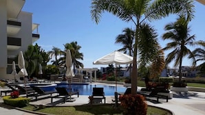 A pool area with lounge chairs and umbrellas, surrounded by palm trees and a building in the background. - Condos SB (Mazatlán)