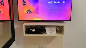 Un televisor montado en la pared con un estante debajo que muestra una caja roja y un dispositivo negro.