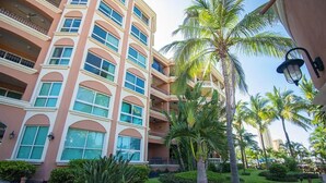 Un hotel de varios pisos con balcones, un jardín cuidado con palmeras y un cielo despejado.