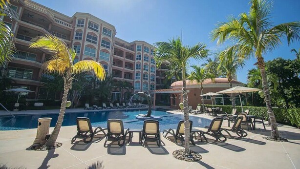 Zona de piscina de hotel con sillas de descanso, palmeras y un edificio grande al fondo.