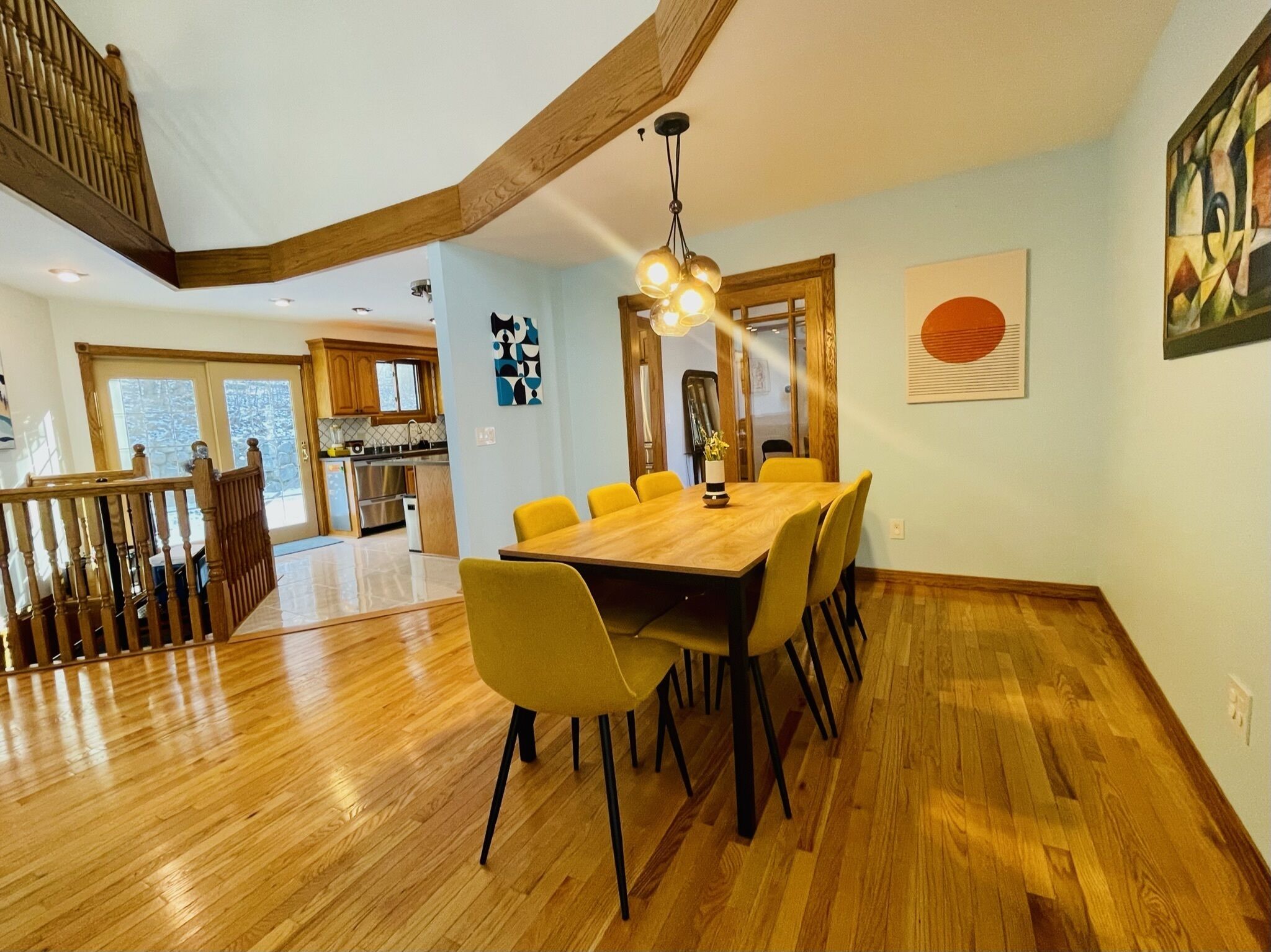 A dining area with a wooden table and yellow chairs, a kitchen in the background, and a staircase leading to an upper level.