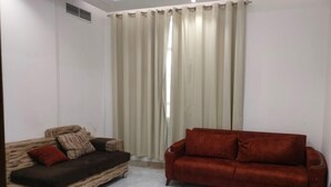 Un salon avec un canapé marron, un canapé rouge et une grande fenêtre ornée de rideaux beige.